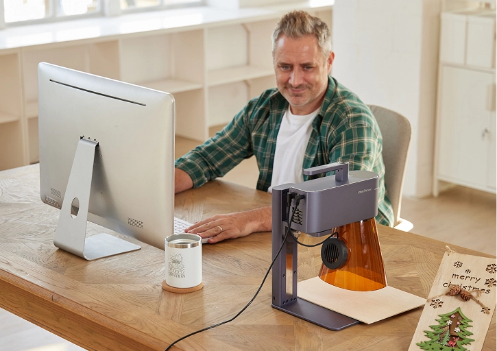 A small business owner using a computer for custom product design while a LaserPecker LP4 engraves stock nearby in a workshop.
